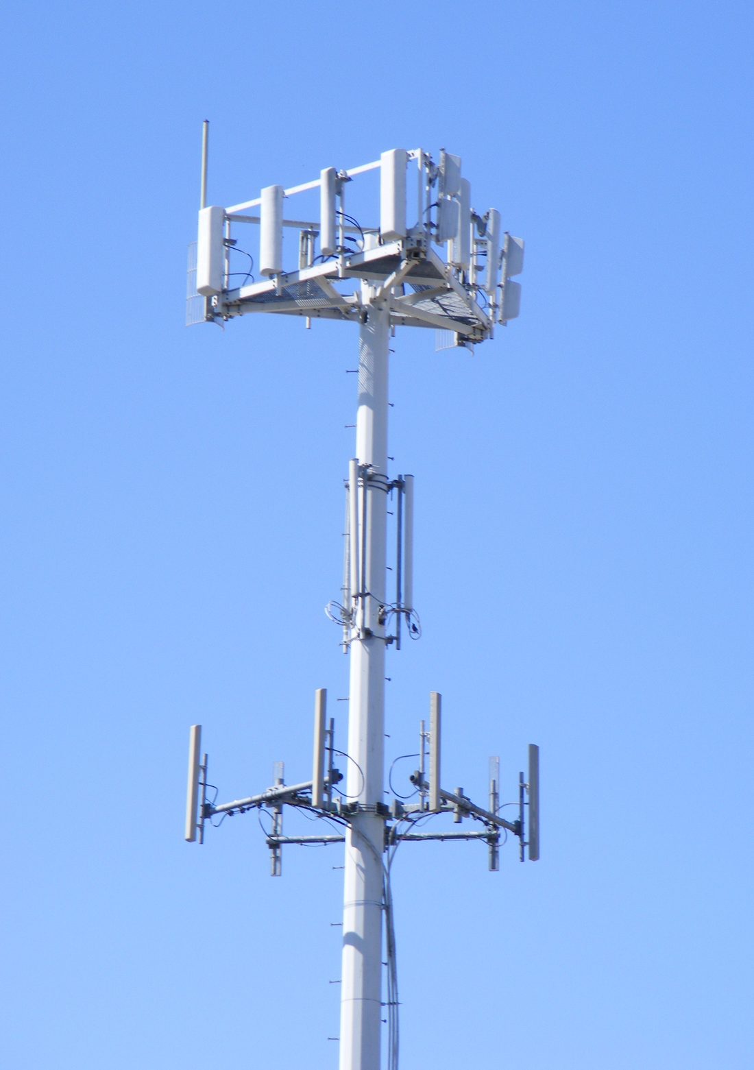 Bir hücresel baz istasyonu kulesinin tepesi: dikdörtgen panel antenler farklı yönlere bakacak biçimde direğe dizilmiş. Her panelin baktığı yön ayrı bir hücre demek; yürürken bağlantın bu panellerden biriyle diğeri arasında devrediliyor.